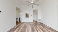 This two-floor space is defined by its striking verticality and clean, minimalist design. The main living area commands attention with soaring ceilings and a ceiling fan positioned at considerable height, reinforcing the open, airy atmosphere. Rustic wood-look plank flooring in warm, varied brown tones runs consistently throughout, providing a cohesive and grounding element across both levels.

The living area features a pass-through window opening to the kitchen, which is equipped with gray cabinetry, granite-style countertops, a white electric range, and a gray subway tile backsplash. A hallway with built-in closet storage connects the main areas and leads to additional rooms, including a bedroom on the upper level that benefits from large multi-pane windows offering generous natural light and exterior views, as well as a vaulted ceiling detail.

The bathroom is finished in an all-white palette, featuring a single-vanity with a mirrored medicine cabinet, and a full soaking tub with an overhead shower and built-in shelving niches. A staircase with black metal railings connects the two floors. Throughout, white walls, recessed vents, and clean architectural lines contribute to a bright, well-organized living environment.