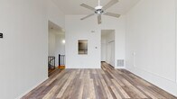 This two-floor space is defined by its striking verticality and clean, minimalist design. The main living area commands attention with soaring ceilings and a ceiling fan positioned at considerable height, reinforcing the open, airy atmosphere. Rustic wood-look plank flooring in warm, varied brown tones runs consistently throughout, providing a cohesive and grounding element across both levels.

The living area features a pass-through window opening to the kitchen, which is equipped with gray cabinetry, granite-style countertops, a white electric range, and a gray subway tile backsplash. A hallway with built-in closet storage connects the main areas and leads to additional rooms, including a bedroom on the upper level that benefits from large multi-pane windows offering generous natural light and exterior views, as well as a vaulted ceiling detail.

The bathroom is finished in an all-white palette, featuring a single-vanity with a mirrored medicine cabinet, and a full soaking tub with an overhead shower and built-in shelving niches. A staircase with black metal railings connects the two floors. Throughout, white walls, recessed vents, and clean architectural lines contribute to a bright, well-organized living environment.