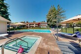 Located in Napa, CA, this well-appointed outdoor amenity area offers a resort-like experience centered around a generously sized swimming pool with crystal-clear blue water. The pool deck is finished in warm concrete and provides ample space for relaxation and recreation. Adjacent to the main pool is a dedicated hot tub/spa, complete with handrail entry and ideal for unwinding after a swim. The surrounding deck is thoughtfully furnished with a variety of seating options, including wicker lounge chairs with plush cushions, cantilever umbrellas for shade, and multiple outdoor dining sets arranged on area rugs — perfect for casual gatherings. A poolside cabana-style structure with large glass windows adds a stylish architectural element and additional shade along the pool's edge. The entire area is enclosed by a black metal security fence with well-maintained green lawn beyond, offering both safety and an attractive boundary. Towering mature trees frame the perimeter, providing natural shade and a sense of privacy. The space enjoys abundant natural light under open California skies, creating a bright and inviting atmosphere year-round. The surrounding residential buildings, with their pitched rooflines and neutral exteriors, complete the community-oriented setting of this outdoor retreat.