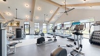 This well-appointed fitness center offers a comprehensive workout environment spread across a single, generously sized floor. The space is defined by its striking vaulted ceiling with exposed wooden beam accents, soaring overhead to create an open, airy atmosphere throughout the main cardio and strength training areas. Large, black-framed windows line the walls, flooding the interior with abundant natural light and offering views of the surrounding outdoor greenery.

The main workout floor is divided into distinct zones. The cardio area features a full lineup of modern treadmills, ellipticals, upright bikes, and recumbent bikes—many equipped with individual screens—all positioned to take advantage of the panoramic window views. The strength training zone is outfitted with a cable machine system, a Smith machine, a dumbbell rack, and various weight benches, all set on durable rubberized flooring.

A separate studio room provides a versatile, open space with light hardwood-style flooring, ceiling fans, and floor-to-ceiling mirrors framed in sleek black metal. This room is equipped with stability balls, medicine balls, kettlebells, and resistance equipment, making it ideal for group fitness, stretching, or functional training. Multiple ceiling fans throughout the facility ensure comfortable airflow, while recessed and pendant lighting complete the clean, modern aesthetic.