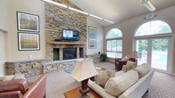 This single-floor clubhouse offers an inviting and well-appointed gathering space designed with both comfort and functionality in mind. The centerpiece of the main lounge is a striking floor-to-ceiling stacked stone fireplace, complete with a mounted flat-screen television and a hearth ledge — creating a warm, focal point that anchors the entire room. Soaring vaulted ceilings with clerestory windows flood the space with natural light, while large arched windows along the exterior wall frame verdant outdoor views and provide a bright, airy atmosphere. The lounge area is generously sized, furnished with plush sofas, leather accent chairs, and a wooden coffee table, all set atop patterned carpeting that adds texture and warmth underfoot. Decorative wall art and diamond-shaped wall accents contribute tasteful visual interest throughout. A separate dining area with a wooden table and chairs offers additional gathering space. The fully equipped kitchen features warm maple cabinetry, stainless steel appliances including a refrigerator, range, and microwave, a tiled floor, and a breakfast bar with pendant lighting — making this space well-suited for hosting events. The open layout and seamless flow between areas make this clubhouse a versatile and welcoming community hub.