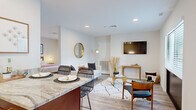 TRELLIS Herndon Unit 2 is a thoughtfully designed one-bedroom, one-bathroom residence that maximizes its compact footprint through an efficient open-concept layout. The single-floor plan seamlessly connects the living, dining, and kitchen areas, creating a cohesive and functional living environment finished with light wood-look flooring throughout.

The living area features recessed lighting, a wall-mounted flat-screen television, and a gold-accented round mirror that adds a modern, decorative touch. The kitchen is a standout feature, boasting white shaker cabinetry with brushed gold hardware, a classic subway tile backsplash, granite countertops, stainless steel appliances, and a dark-base kitchen island with granite surface seating — ideal for casual dining.

The bedroom offers a comfortable, private retreat with a window fitted with blinds that allows natural light, complemented by wall art and hardwood-style flooring. The bathroom includes a granite vanity with white cabinetry and a large mirror, and notably incorporates an in-unit washer for added convenience.

Throughout the unit, the clean white walls and consistent warm-toned finishes create a bright, modern, and cohesive ambiance well-suited for single occupancy living.