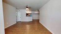 This single-floor space is designed with an open-concept layout that seamlessly connects the living area to the kitchen, creating a sense of spaciousness and ease of movement throughout. The flooring is finished in warm-toned hardwood-style planks that run consistently across the entire space, adding visual continuity and a welcoming aesthetic.

The living area is well-lit by a combination of recessed ceiling lights and a ceiling fan with an integrated light fixture, ensuring both comfort and functionality. Neutral gray-white walls provide a clean, versatile backdrop suitable for a variety of interior styles.

The kitchen is partially open to the main living space, separated by a raised breakfast bar that doubles as a casual dining or workspace surface. The kitchen cabinetry features a warm wood tone, complementing the flooring, and includes built-in appliances such as a microwave. HVAC vents are thoughtfully integrated into the walls and ceiling for climate control.

A doorway visible along one wall suggests access to additional rooms or hallways, and a barn-style sliding door adds a subtle design detail. Overall, the space presents a clean, modern, and functional environment with consistent finishes throughout.