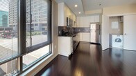 Located in the heart of Hartford, CT, this single-floor unit at 26-44 Front St presents a well-designed layout with contemporary finishes throughout. The kitchen serves as a functional centerpiece, featuring stainless steel appliances including a range, over-the-range microwave, dishwasher, and refrigerator. White cabinetry with clean-lined hardware contrasts sharply against dark granite countertops and a mosaic tile backsplash, while rich, dark hardwood flooring extends across the space, adding warmth and visual depth. Large windows along the kitchen and living areas invite natural light and frame views of the surrounding urban landscape, creating a bright and connected atmosphere. The bedroom offers a quieter retreat, finished with neutral carpet and a window overlooking the neighboring cityscape. The bathroom is well-appointed with large-format gray tile flooring, dark espresso cabinetry, a granite-topped vanity, a framed mirror, and clean white fixtures. The overall design balances modern aesthetics with practical functionality, making this a polished urban residence in downtown Hartford.