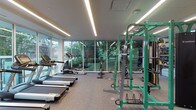 This fitness center offers a thoughtfully designed workout environment that balances functionality with modern aesthetics. The space is anchored by expansive floor-to-ceiling glass walls that flood the interior with natural light and frame views of the surrounding greenery, creating an energizing atmosphere for exercise.

The main workout area is generously proportioned, featuring durable rubber-aggregate flooring suited for high-traffic use. A dedicated cardio zone lines the windowed perimeter, housing multiple treadmills, elliptical machines, stationary bikes, and spin cycles — all positioned to take advantage of the outdoor views. A vibrant green cable and strength training rack serves as a centerpiece for resistance training, complemented by a full dumbbell rack and additional weight equipment.

A warm entryway area features a striking horizontal wood-slat accent wall and open shelving stocked with fresh towels, lending a boutique gym feel. A large motivational wall mural and water fountain station add character near the entry corridor, which also connects to a functional storage and free weights alcove.

Linear LED lighting runs the length of the ceilings, providing consistent illumination throughout. The combination of natural wood tones, clean white walls, and sleek equipment gives the fitness center a polished, contemporary character suited for a comprehensive daily workout routine.