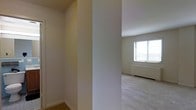 This single-floor residential space offers a clean and functional layout designed with simplicity and comfort in mind. The interior features a neutral color palette throughout, with crisp white walls that lend a bright, airy feel to every room. The living area is finished with wall-to-wall carpeting, providing warmth and a soft underfoot feel, while hard flooring is visible in the transitional hallway areas. Natural light plays a key role in the ambiance of the space, with a large window fitted with horizontal blinds allowing ample daylight to fill the main living area. The architectural detailing is straightforward and practical, with clean-lined walls, standard-height ceilings, and well-defined room separations created through structural columns and partition walls. A closet or storage area is accessible off the hallway, adding useful organizational space. A baseboard heating unit is visible along the wall beneath the window, suggesting efficient climate control integrated into the layout. Overall, the space presents a tidy, move-in-ready environment with a calm, neutral aesthetic suited to a variety of furnishing styles and personal preferences.