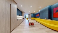 Located at 422 Cherry Street in Toronto's emerging Canary District, this thoughtfully designed single-floor amenity space combines playful energy with refined modern interiors. The layout consists of a welcoming hallway and a generously proportioned living room that together create a cohesive and inviting environment.

The living room is the heart of the space, immediately distinguished by its bold teal accent walls adorned with vibrant, large-format artwork. A signature serpentine yellow sofa anchors the room, offering a lively focal point complemented by a dedicated children's activity area complete with a low table and colorful seating. Light oak hardwood flooring runs throughout, lending warmth and continuity.

A built-in kitchenette, framed by floor-to-ceiling light wood cabinetry, features a sink, microwave, and a marble-style countertop with under-cabinet accent lighting — adding a practical yet polished touch. A wall-mounted flat-screen TV sits above a modular wood cubby unit, with a tall indoor plant adding a natural, organic element.

Toward one end of the space, floor-to-ceiling windows frame open city views and flood the area with natural light, accompanied by a round table and soft seating, ideal for quieter moments. The overall ambiance balances functionality with a bright, welcoming aesthetic suited for residents of all ages.