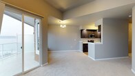 This single-floor space showcases an open-concept layout that seamlessly connects the living area and kitchen into one cohesive environment. The living area is finished with plush, neutral-toned carpeting that adds warmth underfoot, while the walls are painted in a soft gray that lends a clean, contemporary feel throughout.

The kitchen is positioned along the far wall and is defined by a raised breakfast bar peninsula, offering a functional divide between cooking and living spaces. Dark espresso-toned cabinetry provides strong contrast against the light countertops, and the kitchen is equipped with stainless steel appliances, including a microwave and range. The raised ceiling height above the kitchen adds a sense of vertical openness to the space.

A small chandelier with warm bulbs anchors the dining area between the living space and kitchen, providing focused ambient lighting. Natural light flows generously into the room through a full-height sliding glass door on the left wall, which also provides direct access to an outdoor area. The combination of neutral finishes, modern fixtures, and an open floor plan creates a bright and functional living environment.