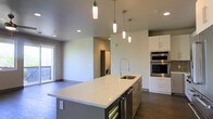This single-floor space showcases a contemporary open-concept design that seamlessly integrates the kitchen and living areas. The kitchen serves as the centerpiece, anchored by a generously sized island topped with striking white quartz countertops featuring subtle veining. The island is thoughtfully equipped with a stainless steel sink, a professional-style pull-down faucet, built-in wine storage, and additional drawers for ample storage.

Overhead, a cluster of sleek cylindrical pendant lights in varying heights adds visual interest and targeted illumination above the island, complementing the recessed lighting that brightens the broader space. The kitchen cabinetry features clean-lined white upper cabinets paired with darker lower island cabinetry, creating a modern two-tone contrast. Stainless steel double wall ovens are integrated into the cabinetry for a polished, professional look.

The open layout flows naturally into the adjacent living area, which benefits from natural light through a window. In the background, a dedicated laundry area is conveniently tucked within the unit. Dark hardwood-style flooring runs throughout, grounding the light-toned walls and creating a cohesive, modern aesthetic.