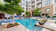 Located at 3521 Wilshire Way in Richardson, Texas, this thoughtfully designed courtyard amenity space is nestled within a modern multi-story residential community. The centerpiece of the outdoor area is a stunning resort-style pool adorned with dramatic water jets and decorative fountain features, framed by mature trees and lush landscaping that lend a sense of natural tranquility to the urban setting. Surrounding the pool, a generous sun deck is lined with cushioned lounge chairs and shaded by navy blue umbrellas, creating an inviting space for relaxation. Vibrant seasonal plantings and sculptural planters add color and texture throughout. A covered outdoor lounge area features exposed white brick walls, stylish sputnik pendant lighting, a wall-mounted flat-screen TV, and a granite-topped bar with counter seating, along with a built-in refrigerator for added convenience. An additional covered social space offers high ceilings, a flat-screen TV, communal high-top tables, and a wall-mounted game board, ideal for casual gatherings. Manicured greenery and ornamental grasses line the walkways, connecting the various zones. The overall ambiance is contemporary and resort-inspired, offering residents a polished and functional outdoor living experience.