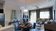 This single-floor space showcases an open-concept layout that seamlessly connects the dining and living areas, creating a cohesive and functional environment. The dining area features a round wooden table accompanied by sleek blue chairs and is illuminated by an elegant pendant light that hangs from the ceiling, adding a warm, inviting focal point to the space.

Adjacent to the dining area, the living room is anchored by a ceiling fan that provides both comfort and style. A large sliding glass door dominates the far wall, allowing abundant natural light to flood the interior while offering views of the outdoor surroundings. Neutral-toned curtains frame the door, adding a sophisticated touch to the room. The living area is further enhanced by a comfortable sofa, a patterned area rug, and a sleek coffee table, all contributing to a modern yet relaxed atmosphere.

The walls are finished in a cool gray tone, complementing the dark hardwood-style flooring that runs throughout. Decorative elements, including wall art and accent pieces, are thoughtfully placed to enhance the overall aesthetic, making this space both visually appealing and highly functional.