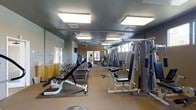 This fitness center offers a spacious, single-floor layout designed to accommodate a wide variety of workout routines. The space is outfitted with an impressive selection of strength training equipment, including a multi-station cable machine, adjustable weight benches, and a full dumbbell rack with weights ranging across multiple increments — ideal for both beginners and experienced lifters. Cardio equipment, including at least one treadmill, is visible toward the rear of the space, providing options for cardiovascular training alongside the strength-focused offerings. Large wall mirrors line portions of the room, enhancing the sense of openness and allowing users to monitor their form during exercises. The ceiling features rows of recessed fluorescent light panels that illuminate the entire floor evenly, ensuring a bright and functional environment. The flooring combines rubber matting in the free-weights area with low-pile carpet throughout the remainder of the space, offering both durability and comfort underfoot. Neutral-toned walls and a clean, organized layout contribute to a focused and motivating atmosphere. The fitness center is thoughtfully arranged to maximize usable floor space while maintaining clear pathways between equipment stations.