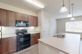 Located at 2001 Wood Hollow Place in Frederick, MD, this single-floor, 2-bedroom, 1-bathroom residence offers a well-designed layout that balances functionality and comfort. The kitchen serves as a central hub of the home, featuring rich medium-toned wood cabinetry, light countertops, and a full suite of black appliances including a range with over-the-range microwave and a refrigerator. A peninsula with an integrated sink and pendant lighting above creates a natural gathering point and provides a seamless transition into the open dining and living area. The living space is bright and airy, with large windows dressed in blinds that invite generous natural light and offer views of the surrounding landscape. Sliding glass doors in one room lead to a balcony, extending the living space outdoors. Both bedrooms are carpeted for warmth and comfort, with the spacious primary bedroom boasting corner windows on two walls for abundant light. The bathroom is clean and functional, featuring a wood-toned vanity with light countertop, a well-lit mirror, and hardwood-style flooring consistent with the main living areas. A dedicated closet and in-unit laundry area with a washer and water heater add practical convenience to this thoughtfully arranged home.