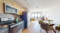 This well-appointed single-floor unit showcases a thoughtfully designed open-concept layout that seamlessly connects the kitchen, dining, and living areas. Light hardwood flooring runs throughout, contributing to a warm and cohesive aesthetic.

The kitchen is outfitted with stainless steel appliances — including a refrigerator, electric range, and built-in microwave — complemented by two-tone cabinetry featuring light upper cabinets and dark lower units, a textured stone tile backsplash, and dark countertops. Track lighting overhead adds both function and style.

The dining area features a round glass-top table paired with mid-century modern upholstered chairs in earthy olive and sage tones. The open living room is bathed in natural light from floor-to-ceiling sliding glass doors that frame expansive panoramic views of the surrounding cityscape and treetops, creating a striking focal point.

The bedroom continues the bright, airy feel with its own large window offering outdoor views, light hardwood floors, and ample space. The full bathroom is finished with marble-look tile flooring, a wood vanity with dark countertop, a backlit mirror, and a tub-shower combination. Throughout, the space maintains a clean, contemporary aesthetic accented by mid-century modern design elements.