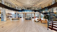 Located at 1000 Vermella Way in New Brunswick, NJ, this expansive amenity floor is an architectural statement in contemporary design. The centerpiece is a breathtaking double-height rotunda lobby, defined by polished large-format tile flooring, cylindrical columns, and a sweeping curved mezzanine level. A sculptural open staircase with sleek black metal railings and glass panels connects the levels, while an elaborate array of suspended pendant lighting creates a dramatic canopy overhead. Floor-to-ceiling glass walls and oversized windows flood the space with natural light and provide views of the surrounding outdoor areas. The lobby transitions seamlessly into distinct social zones, including a lounge area with plush seating anchored by a fireplace and wall-mounted display screen, and a co-working area outfitted with large oval worktables, rolling office chairs in bold red, and branch-inspired pendant fixtures. Additional spaces include a games room featuring a dartboard, a marble-topped dining table, and a well-appointed wine display, as well as a secondary lounge entrance highlighted by veined marble tile flooring and elegant chandelier lighting. Throughout, the design blends warm wood tones, polished stone surfaces, and curated furnishings to create a sophisticated, welcoming environment.