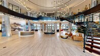 Located at 1000 Vermella Way in New Brunswick, NJ, this expansive amenity floor is an architectural statement in contemporary design. The centerpiece is a breathtaking double-height rotunda lobby, defined by polished large-format tile flooring, cylindrical columns, and a sweeping curved mezzanine level. A sculptural open staircase with sleek black metal railings and glass panels connects the levels, while an elaborate array of suspended pendant lighting creates a dramatic canopy overhead. Floor-to-ceiling glass walls and oversized windows flood the space with natural light and provide views of the surrounding outdoor areas. The lobby transitions seamlessly into distinct social zones, including a lounge area with plush seating anchored by a fireplace and wall-mounted display screen, and a co-working area outfitted with large oval worktables, rolling office chairs in bold red, and branch-inspired pendant fixtures. Additional spaces include a games room featuring a dartboard, a marble-topped dining table, and a well-appointed wine display, as well as a secondary lounge entrance highlighted by veined marble tile flooring and elegant chandelier lighting. Throughout, the design blends warm wood tones, polished stone surfaces, and curated furnishings to create a sophisticated, welcoming environment.