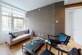 Located at 1277 E 14th St in the Flatbush neighborhood of Brooklyn, NY, this thoughtfully designed residence combines modern aesthetics with functional living. Throughout the space, warm hardwood flooring provides a cohesive foundation, complementing the clean white walls and contemporary design elements.

The kitchen is outfitted with wood-toned cabinetry extending to the ceiling, stainless steel appliances including a French door refrigerator and gas range, a geometric white tile backsplash, and matte black fixtures — delivering both style and practicality.

The living area features a striking chevron-patterned accent wall, large windows that invite natural light, and views of the surrounding Brooklyn streetscape, creating an inviting and airy atmosphere.

The primary bedroom showcases a bold teal geometric accent wall paired with wide windows offering neighborhood views. A second bedroom benefits from a wraparound corner window arrangement, flooding the room with light from multiple directions.

Two full bathrooms reflect a consistent modern design language, each featuring wood-vanities with white countertops, large-format wall tiles, penny tile flooring, and generous mirrors. One bathroom includes a soaking tub with shower, while the other offers a sleek glass-enclosed walk-in shower stall.