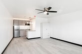 This single-floor unit offers a clean, modern design centered around an open-concept layout that seamlessly integrates the living and kitchen areas. Light-toned hardwood-style flooring runs throughout, lending warmth and continuity to the space, while crisp white walls and bold dark baseboards create a sharp, polished contrast.

The generous living area is highlighted by a ceiling fan with integrated lighting, providing both comfort and illumination. A freestanding white storage cabinet offers practical built-in organization within the main living space.

The kitchen is thoughtfully designed with gray shaker-style cabinetry, granite countertops, and a peninsula that defines the cooking area while remaining open to the living space. Pendant lights hang above the peninsula, adding a contemporary touch. Stainless steel appliances, including a full-size refrigerator and built-in microwave, complete the kitchen. Warm accent lighting above the upper cabinets adds ambiance.

The bathroom is a compact, functional space finished in white, featuring a toilet, ventilation, and what appears to be an in-unit washer and dryer stacked nearby. A window allows natural light into the main living area, enhancing the bright, airy feel of this efficiently designed unit.