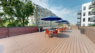 This expansive rooftop deck offers a well-appointed outdoor retreat set atop a modern residential building. The deck is finished with warm-toned composite wood planking that extends generously across the open-air space, providing ample room for both relaxed lounging and outdoor dining. Multiple seating arrangements are thoughtfully distributed throughout the deck, including tables paired with vibrant orange chairs shaded by large navy blue market umbrellas, ideal for al fresco dining or casual gatherings. A comfortable upholstered sofa offers a relaxed lounge area, complemented by a grill station and decorative planters that introduce greenery to the rooftop setting. Horizontal wood-tone privacy panels line portions of the perimeter, creating a sense of enclosure while maintaining an open, airy feel. The surrounding views showcase a mix of classic brick and modern residential architecture, along with a lush mature tree canopy that softens the urban backdrop. A lower-level section of the deck provides additional semi-private space framed by the building's contemporary façade, featuring dark panel cladding and glass-railed balconies above. The rooftop benefits from abundant natural light and open sky exposure, making it a versatile amenity space suited for both social events and quiet outdoor relaxation.