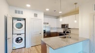 This thoughtfully designed single-floor residence combines modern finishes with functional living. The kitchen serves as a centerpiece, showcasing granite countertops on both the island and perimeter surfaces, rich two-tone cabinetry with white upper cabinets and dark lower cabinets, and a full suite of stainless steel appliances including a French door refrigerator, electric range with microwave, and a built-in oven. A textured tile backsplash adds visual interest, while pendant lighting over the island creates an inviting atmosphere. Light wood-style flooring flows throughout the main living areas, complemented by recessed lighting throughout. A convenient in-unit stacked washer and dryer is neatly tucked into a dedicated laundry alcove. The living area offers generous proportions with a ceiling fan and access to an outdoor balcony, allowing natural light to fill the space. The bedrooms are carpeted for comfort, each featuring a ceiling fan and windows with outdoor views. The bathroom is well-equipped with a dual-sink granite vanity, dark cabinetry with brushed nickel hardware, large wall mirror, and a tub-shower combination with tiled surround. The overall ambiance is clean, bright, and move-in ready.
