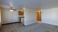 This single-floor unit features an open-concept layout that connects the kitchen and living area, creating a functional and cohesive living space. The kitchen is equipped with warm oak-toned upper cabinetry, a laminate countertop, and a white dishwasher, with tile flooring that transitions seamlessly into the carpeted living area. A half-wall partition subtly defines the kitchen zone while maintaining the open feel of the space. The living area is finished with neutral-toned carpet and includes a ceiling fan, contributing to both comfort and airflow. A textured ceiling runs throughout, adding a subtle design detail overhead. Black baseboard trim provides a clean contrast against the light-colored walls. A hallway visible from the main living space leads to additional rooms, with warm wood-toned doors adding consistency to the interior palette. The overall aesthetic is practical and understated, with a neutral color scheme that offers flexibility for personalization. The layout is straightforward and efficiently organized, making good use of the available space across the single floor.