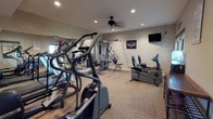 This fitness center is thoughtfully designed to support a comprehensive workout routine. The space is outfitted with a diverse array of exercise equipment, including elliptical machines and cardio steppers positioned along the mirrored wall, which visually expands the room and allows users to monitor their form during workouts. A multi-station cable weight machine anchors the strength training area, providing options for a wide variety of resistance exercises. A recumbent bike offers a low-impact cardio alternative, catering to users of varying fitness levels. The flooring consists of durable, commercial-grade carpet tiles well-suited for high-traffic fitness use. Recessed lighting throughout the ceiling ensures the space is brightly and evenly illuminated, while a ceiling fan promotes air circulation during intense workouts. A wall-mounted flat-screen television adds an entertainment element, making workouts more engaging. A framed print on the wall contributes a personal touch to the otherwise functional environment. The neutral-toned walls and clean layout create an organized, distraction-free atmosphere conducive to focused exercise.