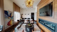 This stylish single-floor lounge space makes a strong architectural statement through its generous ceiling height, which amplifies the sense of openness and grandeur throughout the interior. The most commanding focal point is a large-scale gold sputnik chandelier that anchors the main seating area, casting a warm, sculptural presence overhead. Complementing it are three sleek black pendant lights suspended at varying heights, adding layered illumination across the space.

The feature wall is clad in warm horizontal wood planking, housing a wide-format flat-screen television above a long, linear gas fireplace with a glass front—creating a cohesive and refined entertainment focal point. Dark-stained hardwood floors run throughout, providing a rich contrast to the crisp white walls and light-toned furnishings.

The seating arrangement includes contemporary white and orange accent chairs paired with a round black coffee table atop a geometric-patterned area rug, offering a comfortable gathering area. A high-top bar table with black stools lines the perimeter near floor-to-ceiling sheer-draped windows that welcome abundant natural light and outdoor views. A large abstract canvas painting adds a vibrant artistic touch, while black-framed glass doors connect to adjacent rooms, maintaining visual flow throughout.
