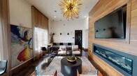 This stylish single-floor lounge space makes a strong architectural statement through its generous ceiling height, which amplifies the sense of openness and grandeur throughout the interior. The most commanding focal point is a large-scale gold sputnik chandelier that anchors the main seating area, casting a warm, sculptural presence overhead. Complementing it are three sleek black pendant lights suspended at varying heights, adding layered illumination across the space.

The feature wall is clad in warm horizontal wood planking, housing a wide-format flat-screen television above a long, linear gas fireplace with a glass front—creating a cohesive and refined entertainment focal point. Dark-stained hardwood floors run throughout, providing a rich contrast to the crisp white walls and light-toned furnishings.

The seating arrangement includes contemporary white and orange accent chairs paired with a round black coffee table atop a geometric-patterned area rug, offering a comfortable gathering area. A high-top bar table with black stools lines the perimeter near floor-to-ceiling sheer-draped windows that welcome abundant natural light and outdoor views. A large abstract canvas painting adds a vibrant artistic touch, while black-framed glass doors connect to adjacent rooms, maintaining visual flow throughout.