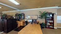 Located at 19321 Park Row Dr in Houston, TX, this single-floor community clubhouse offers a well-appointed and functional space designed for resident comfort and convenience. The interior is finished with warm tan walls, crown molding, and carpeted flooring throughout, creating a cohesive and inviting atmosphere.

The reception and office area serves as the central hub, featuring a large front desk, dual computer workstations, a bulletin board for community notices, and a dedicated café corner equipped with a coffee station and open shelving. Lush artificial palm trees are placed throughout, adding a touch of greenery and warmth to the environment.

Adjacent to the office area, a comfortable lounge provides a relaxed retreat with two leather armchairs and ottomans arranged around a round side table, with a wall-mounted TV overhead. A Wi-Fi sign indicates complimentary connectivity for guests. A large interior mirror expands the visual depth of the space, reflecting views into the main area.

A separate dining and gathering area includes a dining table with chairs, a wall-mounted television, decorative framed artwork, and a classic popcorn machine — ideal for casual socializing. A bookshelf stocked with reading materials rounds out the space, offering a quiet corner for residents to browse. Natural light filters in through multiple windows fitted with white blinds, brightening the overall ambiance.