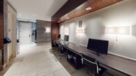 This single-floor business center is thoughtfully designed with a contemporary aesthetic, combining rich dark wood paneling, textured gray wall tiles, and light wood-look porcelain flooring throughout. The space is organized into distinct functional zones that serve the practical needs of its users.

A long, sleek workstation counter runs along one wall, accommodating multiple desktop computers and ergonomic task chairs, making it well-suited for productivity. Wall-mounted sconces provide warm, focused lighting above the workstations, complemented by recessed ceiling lights that illuminate the space evenly.

Adjacent to the workstations, a dedicated refreshment counter is finished in dark wood cabinetry with a deep countertop, featuring a coffee machine, a water dispenser, a utility sink with a brass-finish faucet, and built-in storage below. A digital package management kiosk is mounted near the entrance to a secured package room, streamlining delivery retrieval. The keycard-access entry door, framed in a light stone surround, adds a layer of security. The overall ambiance is polished and professional, with a cohesive material palette that balances functionality with refined, modern design.