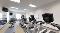 The fitness center is a bright, well-maintained space designed to support a comprehensive workout experience. The room features a drop ceiling with recessed panel lighting that ensures consistent, even illumination throughout. Light-toned walls and neutral flooring contribute to a clean, focused atmosphere.

The cardio section is prominently featured, with a row of TRUE-brand treadmills and elliptical machines aligned to maximize the use of floor space. A large window along one wall allows natural light to filter in, enhancing the open feel of the room. Framed artwork is displayed on the walls, adding a modest decorative touch to the functional environment.

Along the far wall, a dumbbell rack holds a range of free weights, accompanied by a wall-mounted workout guide for reference. Additional strength and cable equipment is visible in the background, rounding out the available training options.

The layout is thoughtfully organized, providing clear pathways between equipment zones for safe and efficient movement. Overall, the fitness center offers a well-rounded, inviting environment suited for both cardio and strength training.