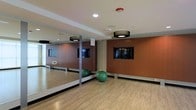 This single-floor fitness studio is thoughtfully designed to support a range of exercise activities. The room features a full-length mirrored wall along one side, enhancing the sense of space and allowing users to monitor their form during workouts. The flooring is a smooth, light-toned hardwood-style surface, providing a clean and supportive foundation suitable for yoga, stretching, cardio, and strength training.

The warm, rich brown accent wall adds a welcoming visual depth to the space and serves as a backdrop for a wall-mounted flat-screen television, ideal for following workout programs or streaming fitness classes. Recessed ceiling speakers suggest built-in audio capabilities. Overhead, modern drum-shade pendant light fixtures provide even, ambient illumination throughout the room.

Exercise equipment includes stability balls, indicating the studio is set up for functional fitness and core training. The ceiling is finished with recessed lighting and integrated safety features, including a fire suppression sprinkler. The overall design balances practicality with a clean, modern aesthetic, creating an inviting and functional environment for individual or group fitness sessions.