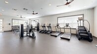 Located at 1851 Encantado Way in Kissimmee, Florida, this fitness center offers a well-organized and fully equipped workout facility on a single level. The space features a clean, contemporary design with white shiplap-style wall paneling, recessed lighting, and multiple ceiling fans that ensure comfortable air circulation throughout. Large windows fitted with blinds allow natural light to fill the room while maintaining privacy, creating a bright and energizing atmosphere.

The cardio zone is prominently featured, housing multiple treadmills and elliptical machines arranged in a row for easy access. Nearby, a multi-station cable machine and additional strength training equipment provide options for targeted resistance workouts. A dedicated free weights area includes a tiered dumbbell rack with a full range of weights, accompanied by adjustable weight benches and full-length mirrors that allow users to monitor their form. Additional weight machines are positioned throughout the floor to maximize the use of available space.

Near the entrance, built-in wooden shelving and storage cabinetry provide a functional and organized area, complemented by a teal accent wall that adds a touch of color to the otherwise neutral palette. The rubber flooring throughout the main workout area offers both durability and comfort underfoot. Overall, this fitness center delivers a comprehensive and welcoming environment suited for a wide variety of exercise routines.