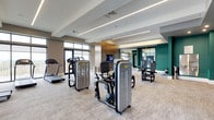 This fitness center offers a spacious, thoughtfully designed environment suited for a comprehensive workout routine. The open floor plan accommodates a generous selection of professional-grade equipment, including strength training machines and cardio equipment such as treadmills, all arranged to allow comfortable movement throughout the space.

The interior design strikes a balance between functional and contemporary aesthetics. Neutral-toned carpeting covers the floor, providing both comfort and durability underfoot. The ceiling features recessed linear lighting that delivers consistent, bright illumination across the entire space. Architectural ceiling details, including tiered tray elements, add visual depth and dimension to the room.

The color palette combines warm beige and taupe wall tones with a bold teal accent wall, creating an energetic yet composed ambiance. Large windows along one wall invite natural light and provide views to the exterior. Mounted televisions are positioned for visibility from multiple workout stations. Full-length mirrors along one section of the space enhance the sense of openness while serving a practical function for form and technique monitoring during exercise.