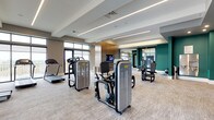 This fitness center offers a spacious, thoughtfully designed environment suited for a comprehensive workout routine. The open floor plan accommodates a generous selection of professional-grade equipment, including strength training machines and cardio equipment such as treadmills, all arranged to allow comfortable movement throughout the space.

The interior design strikes a balance between functional and contemporary aesthetics. Neutral-toned carpeting covers the floor, providing both comfort and durability underfoot. The ceiling features recessed linear lighting that delivers consistent, bright illumination across the entire space. Architectural ceiling details, including tiered tray elements, add visual depth and dimension to the room.

The color palette combines warm beige and taupe wall tones with a bold teal accent wall, creating an energetic yet composed ambiance. Large windows along one wall invite natural light and provide views to the exterior. Mounted televisions are positioned for visibility from multiple workout stations. Full-length mirrors along one section of the space enhance the sense of openness while serving a practical function for form and technique monitoring during exercise.
