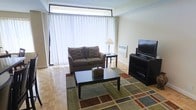 This single-floor space offers a comfortable and functional layout designed to maximize both utility and livability. The living area serves as the centerpiece of the space, featuring warm parquet hardwood flooring that adds a classic character to the interior. Large windows dressed in floor-to-ceiling sheer white curtains allow abundant natural light to filter through, creating a bright and airy atmosphere throughout the day. A sliding glass door provides access to the outdoors, further enhancing the sense of openness. The living room is thoughtfully arranged with a loveseat sofa accented with decorative pillows, a dark wood coffee table set atop a multicolored area rug, and a matching dark wood media console that houses a flat-screen television. A side table with a traditional lamp adds a warm, ambient glow to the space. A baseboard heating unit runs along the exterior wall, ensuring year-round comfort. The neutral color palette of soft whites and warm beiges throughout the walls and furnishings creates a calm, welcoming environment. Overall, this space presents a practical yet inviting atmosphere well-suited for everyday living.