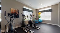 Located at 3785 Codet Rd. in Columbus, Ohio, this fitness center offers a comprehensive, single-floor workout facility designed to support a wide range of exercise routines. The space is generously proportioned, with high ceilings, recessed lighting, and a ceiling fan that contribute to a comfortable and airy atmosphere throughout.

The main cardio area features a row of treadmills equipped with individual display screens, complemented by multiple elliptical cross-trainers positioned along the perimeter. Large windows dressed with decorative valances and horizontal blinds line the walls, allowing abundant natural light to fill the space while providing views of the surrounding exterior. Wall-mounted flat-screen televisions are strategically placed for convenient viewing during workouts.

Full-length mirrors are installed along one wall, enhancing the sense of openness and allowing users to monitor their form. A dedicated strength training zone includes a multi-function cable machine and an adjustable weight bench, while a separate functional training corner houses stability balls, resistance bands, and medicine balls on a rack. The durable rubber flooring throughout provides both safety and comfort underfoot.

Glass-paneled doors offer a view into an adjacent area, suggesting additional amenity spaces nearby. The neutral warm-toned walls and well-organized layout create a clean, motivating environment suited for daily fitness use.