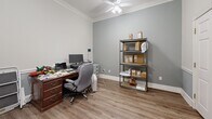 Located at 2900 Baby Ruth Ln in Nashville, Tennessee, this single-room office is a well-appointed and functional workspace with notable architectural detailing. The room features warm hardwood-style flooring throughout, complemented by soft gray-toned walls and crisp white trim work. Elegant crown molding lines the ceiling, while a chair rail with wainscoting adds a classic, refined character to the walls.

A ceiling fan is centrally mounted, providing both light and airflow to the space. The office is equipped with a traditional wood executive desk paired with a rolling office chair, alongside a multi-drawer filing cabinet and a metal shelving unit offering ample storage. A desktop computer and printer setup makes the space fully functional for administrative use.

The room benefits from natural light through a window fitted with horizontal blinds, and a French door with glass panes opens to an adjacent area, enhancing the sense of openness. A decorative six-panel interior door with ornate door casing provides a polished entryway. A small table and chair set near the French door and window adds a versatile secondary work or meeting area. The overall ambiance is professional yet warm, blending classic architectural details with practical office functionality.