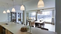 This thoughtfully designed single-floor space showcases a bright, open-concept layout that effortlessly connects the kitchen, dining, and living areas into one cohesive environment. The kitchen features a generous island with a clean, light-toned countertop, ideal for casual gathering or food preparation, while elegant pendant lights hang overhead, adding both warmth and a modern aesthetic. The dining area accommodates a large rectangular table surrounded by upholstered chairs, positioned to take full advantage of the expansive floor-to-ceiling windows that frame sweeping outdoor views. Natural light floods the interior, creating an airy and welcoming atmosphere throughout. The living area is comfortably appointed with soft seating and ambient lighting, offering a relaxed retreat within the open floor plan. Structural columns subtly define each zone without disrupting the flow of the space. The neutral color palette — featuring whites, grays, and warm wood tones — contributes to a refined yet inviting ambiance. The large glazed facade not only maximizes daylight but also visually connects the interior to the surrounding outdoor landscape, making this a versatile and contemporary space well-suited for both everyday living and entertaining.