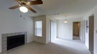 This single-floor residence offers an open and airy layout with a seamless flow between the living and dining areas. The living room is anchored by a tile-surround fireplace, adding both a functional and decorative focal point to the space. Soft, neutral carpeting extends throughout, contributing to a warm and comfortable atmosphere. A ceiling fan with a light fixture keeps the living area well-ventilated, while a classic pendant light illuminates the dining area, adding a touch of understated elegance. Natural light filters in through a window with blinds and a set of vertical-blind-covered sliding glass doors, brightening the interior and providing an easy connection to the outdoors. The textured ceiling adds subtle dimension to the space. The open layout allows for flexible furniture arrangements and creates a sense of spaciousness. A hallway visible in the background hints at additional rooms beyond the main living area, suggesting a well-organized and practical floor plan. Overall, the space presents a clean, neutral palette that provides an excellent canvas for personalization.