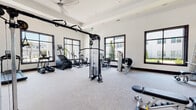 This modern fitness center is thoughtfully designed to support a full range of workout routines within a bright, open, single-floor layout. The space is divided into distinct functional zones, seamlessly connected through glass-paneled doors and an entry area finished with light hardwood flooring. The main workout areas are carpeted for comfort underfoot and feature crisp white walls accented by bold dark window trim and dark baseboards, lending a contemporary, polished aesthetic throughout.

The cardio zone is lined with large, black-framed windows that frame outdoor views of greenery and neighboring structures, flooding the space with natural light. Equipment includes treadmills, elliptical machines, a stair climber, and a stationary bike, all from TRUE Fitness. A ceiling fan overhead ensures comfortable airflow during workouts.

The strength training area is equally well-appointed, offering a cable cross machine, a functional trainer, a multi-station cable pulley system, adjustable benches, a dumbbell rack, and various selectorized strength machines. Full-length mirrors along one wall allow users to monitor their form. Recessed lighting and a tray ceiling add an architectural refinement to this section.

The entry corridor features light hardwood flooring, open shelving, restroom access, and a sanitation station with dispensers and waste receptacles, prioritizing cleanliness and convenience for all users.