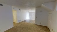 This two-floor residence features a spacious main living area that combines comfort and functionality. The open layout transitions between carpeted and tile flooring, clearly delineating the living and dining zones. White walls throughout create a clean, neutral backdrop, while the textured popcorn ceiling adds a classic architectural detail. A large sliding glass door fitted with vertical blinds serves as the focal point of the room, allowing ample natural light to fill the space and providing access to the outdoors. The flush-mount ceiling light offers practical illumination, complementing the natural light. Integrated HVAC vents in both the ceiling and lower wall areas ensure efficient climate control throughout the space. A doorway visible along one wall leads to additional rooms, hinting at the connectivity of the floor plan. The layout is well-suited for both everyday living and casual entertaining, with the distinct flooring materials helping to define separate functional areas within the open space. The two-floor design offers additional living space beyond the main level, providing separation between living and private areas.