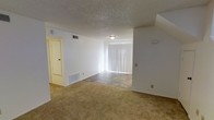 This two-floor residence features a spacious main living area that combines comfort and functionality. The open layout transitions between carpeted and tile flooring, clearly delineating the living and dining zones. White walls throughout create a clean, neutral backdrop, while the textured popcorn ceiling adds a classic architectural detail. A large sliding glass door fitted with vertical blinds serves as the focal point of the room, allowing ample natural light to fill the space and providing access to the outdoors. The flush-mount ceiling light offers practical illumination, complementing the natural light. Integrated HVAC vents in both the ceiling and lower wall areas ensure efficient climate control throughout the space. A doorway visible along one wall leads to additional rooms, hinting at the connectivity of the floor plan. The layout is well-suited for both everyday living and casual entertaining, with the distinct flooring materials helping to define separate functional areas within the open space. The two-floor design offers additional living space beyond the main level, providing separation between living and private areas.