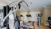 This dedicated home fitness space is thoughtfully designed to support a comprehensive workout routine. The room features light hardwood flooring that adds warmth to the functional environment, while crown molding and recessed ceiling lights lend a polished, finished look that sets it apart from a typical home gym. A large wall mirror spans one side of the room, enhancing the sense of space and allowing users to monitor their form during exercise. The ceiling incorporates an access panel, adding a practical element to the well-maintained interior. The gym is outfitted with an impressive array of equipment, including a multi-station cable and strength training machine, a motorized treadmill with a digital display console, and an elliptical cross-trainer, providing options for both cardiovascular and resistance-based workouts. White paneled doors offer access to additional storage or adjoining spaces, keeping the room organized and uncluttered. Natural light filters in through a window fitted with blinds, complementing the recessed lighting to create a bright and motivating atmosphere. Overall, this space delivers a fully functional, private fitness environment with the comfort and aesthetic detail of a well-appointed interior room.