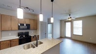This single-story space showcases an open-concept layout that connects the kitchen and living area in a cohesive, functional design. The kitchen serves as a focal point, featuring a granite-topped island with a double stainless steel undermount sink and elegant pendant lighting overhead. A track lighting system provides additional illumination, while warm wood-toned cabinetry offers ample storage. Black appliances, including a range and over-the-range microwave, complement the kitchen's contemporary aesthetic. A tile backsplash adds a clean, finished detail to the cooking area.

The living space extends naturally from the kitchen, finished with hardwood-style flooring that runs throughout. A ceiling fan with a light fixture anchors the living area, contributing to both comfort and style. A full-length glass door floods the space with natural light and provides access to an outdoor area. A doorway visible from the kitchen leads to an additional room, suggesting a flexible floor plan. The neutral color palette and modern finishes throughout create a bright, welcoming ambiance well-suited for everyday living.