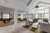 Located at 1321 Lovvorn Rd in Carrollton, Georgia, this single-story community clubhouse offers a generous, open layout designed for both relaxation and social engagement. The centerpiece of the space is a dramatic vaulted ceiling in the main lounge area, which soars overhead and is flanked by tall, multi-pane windows that flood the interior with natural light and offer views of the surrounding greenery and pool area. Wood-look plank flooring laid in a herringbone pattern runs throughout the main corridor and lounge, lending warmth and a modern aesthetic to the bright, white-painted interior. Bold cobalt blue accent walls add a striking pop of color, each framing a wall-mounted television to create comfortable viewing areas within the seating clusters. The lounge is furnished with contemporary gray armchairs, ottomans, and modular sofas arranged across patterned area rugs, creating inviting conversation zones. A long white bar-height communal table with black swivel stools serves as a versatile workspace or social gathering spot, positioned adjacent to a beverage station with a tiled backsplash. The dedicated game room features a pool table, shuffleboard table, patterned carpet, and a mounted television, providing an entertainment-focused retreat. Tall windows throughout ensure the entire space remains bright and connected to the outdoors.