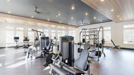 This expansive fitness center is thoughtfully designed to support a full range of workout routines in a bright, modern environment. The space features two distinct ceiling treatments — a sleek dark tray ceiling with recessed lighting and ceiling fans over the strength training area, and a warm wood-paneled vaulted ceiling that crowns the cardio zone, adding architectural character and a sense of openness. Large windows fitted with sheer roller shades line multiple walls, flooding the interior with natural light while maintaining privacy.

The strength training section is stocked with a comprehensive selection of True Fitness cable and selectorized machines, a multi-use power rack with free weights and barbells, adjustable benches, and a dumbbell rack. A large functional training tire anchors the open floor space at the center of the room, ideal for dynamic and cross-training exercises. The cardio area is well-appointed with treadmills, ellipticals, and stationary bikes, each equipped with individual screens. Dark hardwood-style flooring runs throughout, providing a polished yet durable surface. An additional room near the entrance features a small refrigeration unit and a barber-style chair, offering a utility or grooming space. The overall ambiance is clean, spacious, and professional.