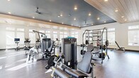 This expansive fitness center is thoughtfully designed to support a full range of workout routines in a bright, modern environment. The space features two distinct ceiling treatments — a sleek dark tray ceiling with recessed lighting and ceiling fans over the strength training area, and a warm wood-paneled vaulted ceiling that crowns the cardio zone, adding architectural character and a sense of openness. Large windows fitted with sheer roller shades line multiple walls, flooding the interior with natural light while maintaining privacy.

The strength training section is stocked with a comprehensive selection of True Fitness cable and selectorized machines, a multi-use power rack with free weights and barbells, adjustable benches, and a dumbbell rack. A large functional training tire anchors the open floor space at the center of the room, ideal for dynamic and cross-training exercises. The cardio area is well-appointed with treadmills, ellipticals, and stationary bikes, each equipped with individual screens. Dark hardwood-style flooring runs throughout, providing a polished yet durable surface. An additional room near the entrance features a small refrigeration unit and a barber-style chair, offering a utility or grooming space. The overall ambiance is clean, spacious, and professional.