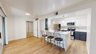 Located at 4963–4967 Owens Dr in Pleasanton, California, this single-floor residence combines contemporary updates with a clean, functional layout. Light-toned hardwood-style flooring flows throughout the main living areas, contributing to a bright and cohesive aesthetic. The open-concept kitchen serves as a central hub, showcasing white upper cabinetry paired with contrasting charcoal lower cabinets, gold hardware accents, and a striking chevron-patterned tile backsplash. A large island with marble-look countertops and pendant lighting overhead offers seating for several bar-height chairs, making it ideal for casual dining and entertaining. Stainless steel appliances, including a refrigerator, range, and microwave, complete the kitchen. The living area is generously proportioned, featuring a ceiling fan, natural light from windows with blinds, and sliding glass door access. The bedroom is carpeted for comfort, includes crown molding, a ceiling fan, a window for natural light, and direct access to a dedicated walk-in closet with built-in shelving. The bathroom features a white vanity with marble-look countertop, gold fixtures, wood-look flooring, a dark accent wall, and a shower-tub combination.
