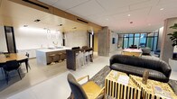 This single-floor space is a masterclass in modern, upscale design, seamlessly combining social, recreational, and lounge functions in one expansive open-plan layout. The interior is anchored by a sleek wet bar and kitchen area, featuring a long white countertop with pendant lighting in a distinctive looped design, complemented by warm wood cabinetry and high-end fixtures. A dark wood bar-height table with white modern barstools serves as a central gathering point, ideal for casual dining or conversation. The flooring throughout is a clean, large-format light tile that enhances the sense of openness and continuity across the space. Exposed concrete columns add an industrial edge that contrasts beautifully with the refined wood and white paneling on the walls and ceiling. The lounge area is furnished with a bold, tufted black leather sofa, sculptural gold-accented side tables, and a plush area rug, creating an inviting and sophisticated seating vignette. Toward the far end of the space, floor-to-ceiling steel-framed glass partitions allow natural light to filter through while framing a vibrant pink billiard table, adding a playful focal point. Recessed lighting throughout ensures the space is well-lit while maintaining an elegant ambiance.